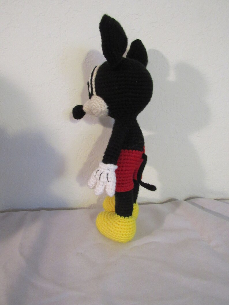 May include: A black and white crocheted Mickey Mouse doll with red shorts and yellow shoes. The doll is standing on a white surface.