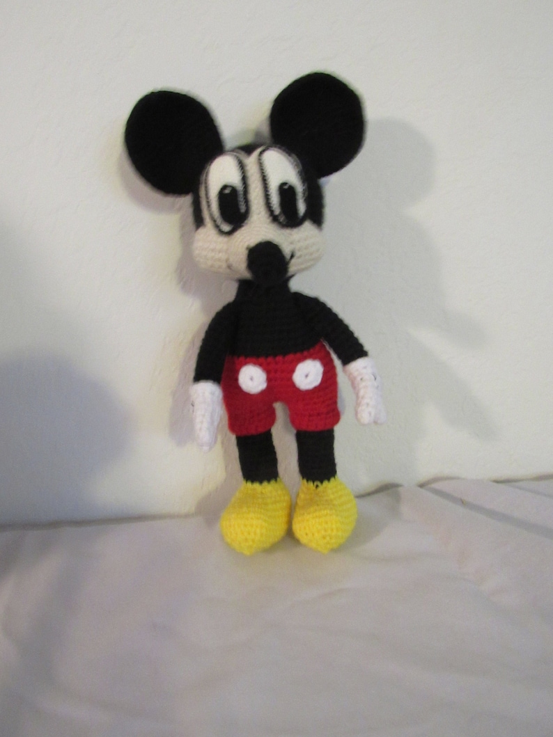 May include: A crocheted plush toy of Mickey Mouse. The toy is black with red shorts and yellow shoes. The toy has large black ears and a white face with black eyes.