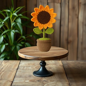 May include: A crocheted sunflower in a brown pot sits on a wooden cake stand with a black base.
