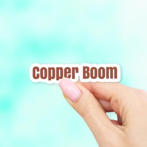 May include: A white sticker with the text "copper Boom" in brown letters. A hand is holding the sticker.
