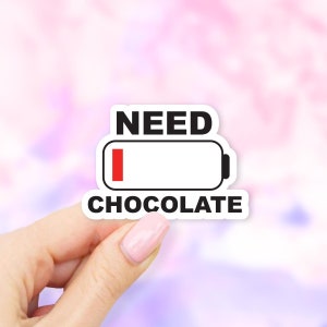 May include: A white sticker with black text that reads "NEED CHOCOLATE" above a battery icon with a red bar indicating low power. The sticker is held by a hand against a soft, colorful background.