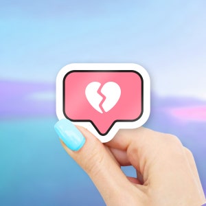 May include: A pink speech bubble sticker with a white broken heart graphic. The sticker is held by a hand with a light blue painted fingernail against a blurred blue and purple background.