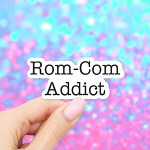 May include: A white sticker with the words "Rom-Com Addict" in black, held by a hand with a pink nail. The background is a blurred mix of blue, pink, and white colors.
