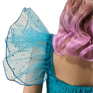 Pode incluir: Um vestido turquesa com uma manga de tule azul com babados e brilho. O vestido tem um decote quadrado e um corpete justo.