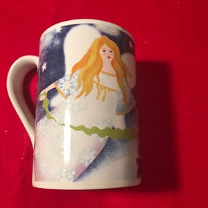May include: White ceramic mug with a blue and white angel design. The angel has long blonde hair and is wearing a white dress with a green sash. The background is a dark blue with white stars.
