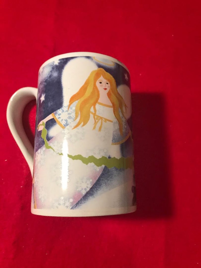 May include: White ceramic mug with a blue and white angel design. The angel has long blonde hair and is wearing a white dress with a green sash. The background is a dark blue with white stars.