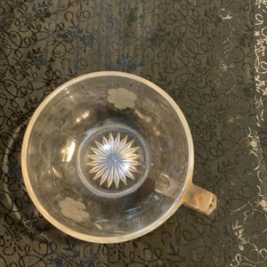 May include: A clear glass teacup featuring a starburst pattern etched into the center of the base and a series of floral motifs engraved around the interior walls. The cup includes a single handle extending from the side of the circular rim.