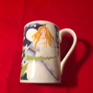 May include: White ceramic mug with a colorful illustration of an angel with blonde hair and a blue dress. The angel is surrounded by a blue and green background with stars.