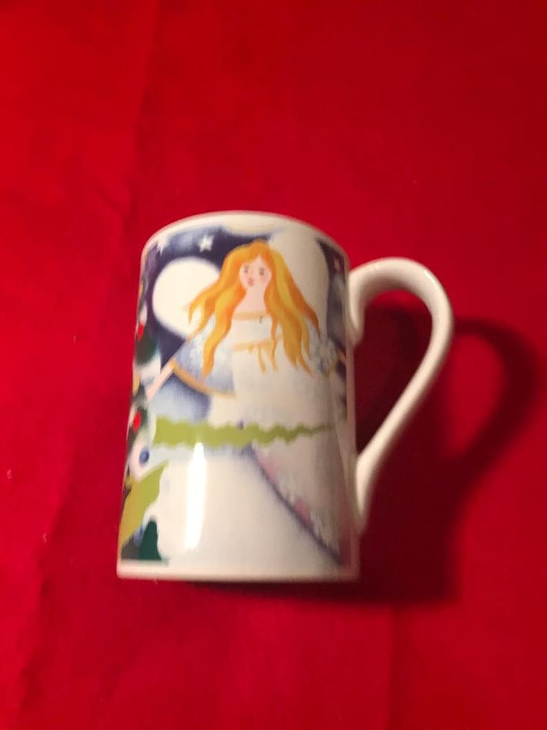 May include: White ceramic mug with a colorful illustration of an angel with blonde hair and a blue dress. The angel is surrounded by a blue and green background with stars.