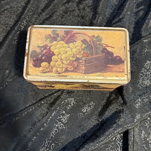 May include: A rectangular metal tin with a vintage fruit illustration. The artwork depicts a basket overflowing with green and purple grapes. The tin has a gold and brown color scheme with a slightly distressed appearance.