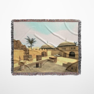 May include: A woven throw blanket featuring a digital illustration of a desert scene with buildings and crates. The scene is reminiscent of a video game map.