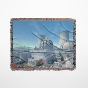 May include: A woven throw blanket featuring a detailed illustration of a nuclear power plant under a blue sky. The design includes cooling towers and other architectural elements. The blanket has a fringed edge.