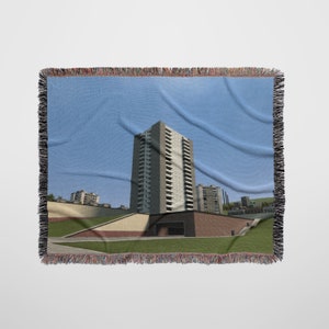 May include: A woven throw blanket featuring a photograph of a tall, modern apartment building with a blue sky in the background.