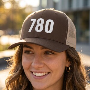 May include: A brown trucker hat with a white "780" graphic on the front. The hat has a mesh back and a curved brim. The hat is worn by a person with curly brown hair.