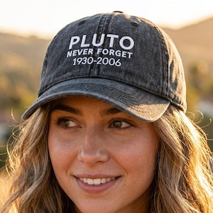 May include: A gray baseball cap with the text "PLUTO NEVER FORGET 1930-2006" in white. The cap has a curved brim and a classic six-panel design. The hat is worn by a person with long, wavy hair.