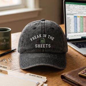 May include: A dark gray baseball cap with the text "FREAK IN THE SHEETS" embroidered in white. The cap is on a wooden desk with a laptop, mug, and folders labeled "TAX PREP 2024" and "CLIENT DATA".