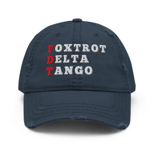 May include: Navy blue distressed baseball cap with the embroidered text "FOXTROT DELTA TANGO" in white and red. The hat has a pre-curved peak and a worn, vintage look.