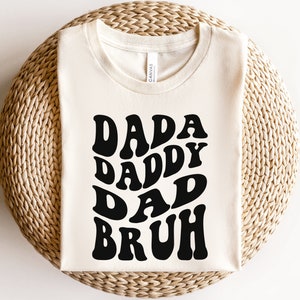 May include: A white t-shirt with a black graphic that says "Dada Daddy Dad Bruh" in a retro font.