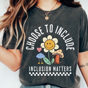 May include: A dark gray t-shirt with a white graphic design that says "Choose to Include" with a smiling flower, a cloud, and a mushroom. The text "Inclusion Matters" is below the graphic.