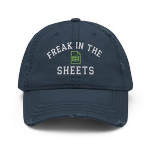May include: A distressed navy blue baseball cap with the text "FREAK IN THE SHEETS" embroidered in white. A green spreadsheet icon is in the center of the text. The cap has a pre-worn look.