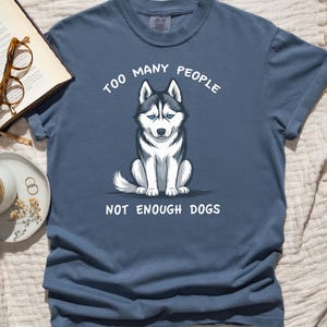 May include: A blue t-shirt featuring the text "TOO MANY PEOPLE NOT ENOUGH DOGS" and a husky dog graphic. The dog is black, white, and grey with blue eyes. The shirt is short-sleeved with a crew neck.