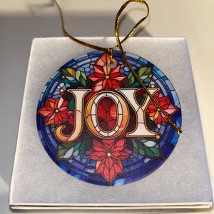 May include: A round ceramic ornament featuring the word JOY in large white letters. The design mimics stained glass with red poinsettia flowers, green leaves, and blue geometric patterns. A thin gold string is attached for hanging purposes.