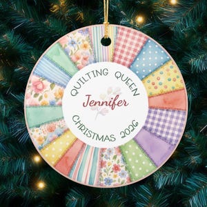 May include: Round Christmas ornament with a patchwork quilt design in pastel colors. The ornament features the text "Quilting Queen Jennifer Christmas 2026" in a circular arrangement around a floral graphic. A gold string is attached for hanging.