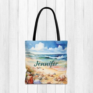 May include: A blue and white tote bag with a watercolor painting of a beach scene and the name "Jennifer" printed on the front. The bag has black handles.