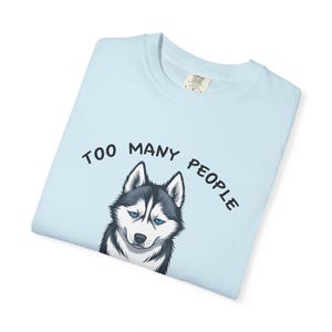 May include: Pale blue t-shirt with a graphic of a husky dog with blue eyes. The text "TOO MANY PEOPLE" is arched above the dog's head in black letters. The shirt is folded and has a tag.