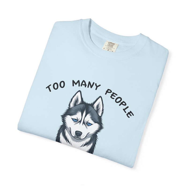 May include: Pale blue t-shirt with a graphic of a husky dog with blue eyes. The text "TOO MANY PEOPLE" is arched above the dog's head in black letters. The shirt is folded and has a tag.