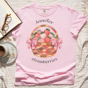May include: A light pink t-shirt featuring a watercolor illustration of a basket filled with strawberries. The basket has a woven design and is adorned with pink bows. The text "Jennifer strawberries" is arched above and below the basket.