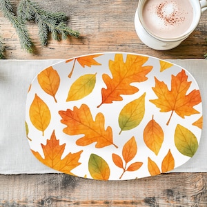 May include: An oval-shaped serving platter with a white background and a pattern of autumn leaves in shades of orange, yellow, and green. The platter is on a white linen runner, with a cup of coffee and pine needles in the background.