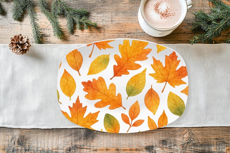 May include: An oval-shaped serving platter with a white background and a pattern of autumn leaves in shades of orange, yellow, and green. The platter is on a white linen runner, with a cup of coffee and pine needles in the background.