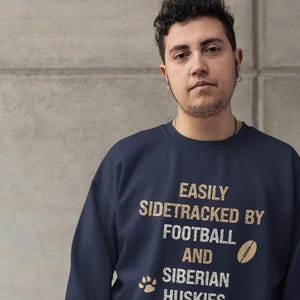 May include: A navy blue sweatshirt with the text "EASILY SIDETRACKED BY FOOTBALL AND SIBIRIAN HUSKIES" in a distressed, gold-colored font. A football and paw print are also included in the design.
