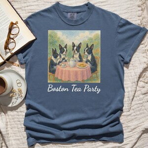 May include: A blue t-shirt features a square graphic of Boston Terriers at a tea party. The dogs are black and white, gathered around a pink table with tea and cookies. The words "Boston Tea Party" are printed below the graphic.