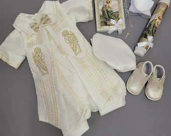 Puede incluir: Un conjunto de bautizo blanco con un pelele con bordados dorados y una pajarita a juego. El conjunto incluye una imagen religiosa enmarcada, una vela, un rosario, una concha, un gorro y un par de zapatos blancos.