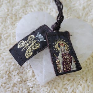 May include: Two small, rectangular religious pendants with embroidered designs and a dark brown cord. One pendant features gold lettering and a crown, while the other depicts a religious figure with a halo and crown.