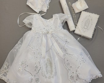 May include: White christening gown with lace and sequins, a white satin bonnet, a white candle, a white rosary, and a white prayer book with an embroidered image of the Virgin Mary.