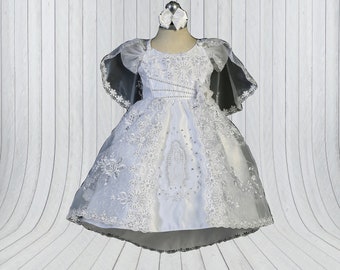 May include: White satin baby dress with lace overlay and a beaded design. The dress features a cape with lace trim and a small flower embellishment. The dress has a picture of the Virgin Mary on the front.