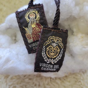 Pode incluir: Dois pequenos pingentes religiosos retangulares com tecido marrom escuro e bordados dourados. Um apresenta a imagem de um santo, o outro um brasão com o texto "VIRGEN DEL CARMEN". Ambos têm cordões marrons escuros.
