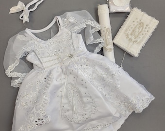 May include: White christening gown with lace and sequins, a white headband, a white candle, a white prayer book, and a white veil. The gown features a design of the Virgin Mary.