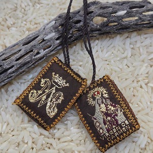 May include: A brown fabric scapular featuring two rectangular panels connected by a dark brown cord. One panel displays a gold embroidered crown and monogram, while the other depicts a figure with the text VIRGEN DEL CARMEN in gold thread.