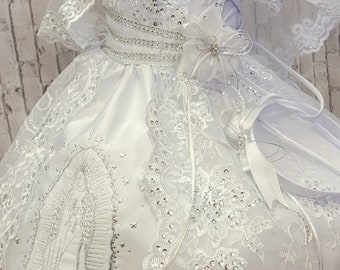 May include: White satin dress with lace and rhinestone embellishments. The dress features a silver embroidered image of the Virgin Mary and stars. The dress comes with a matching lace cape.