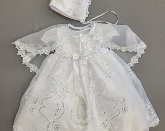 May include: White christening gown with a lace overlay and a matching bonnet. The gown features a silver embroidered image of the Virgin Mary.