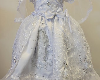 May include: White satin and lace dress with a silver embroidered image of the Virgin Mary. The dress has a tulle overlay and a white satin bow.