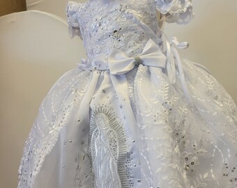 May include: White lace dress with a silver embroidered image of the Virgin Mary. The dress has a satin sash with a bow and a matching headband.