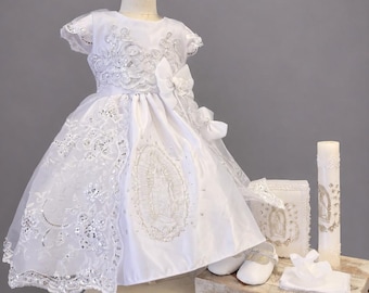 May include: A white dress with short sleeves, featuring intricate silver embroidery, sequins, and a bow at the waist. The skirt displays a central embroidered image of the Virgin of Guadalupe. Includes matching white shoes, a book, and a candle.