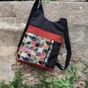Può includere: Borsa a tracolla nera con un accento rosso e un motivo geometrico. La borsa è dotata di una tasca con cerniera e di una lunga tracolla nera da indossare a spalla. Il motivo geometrico comprende triangoli e semicerchi di vari colori.