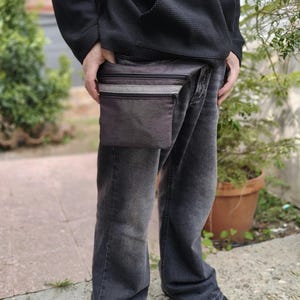 Puede incluir: Un bolso bandolera gris oscuro con dos compartimentos horizontales con cremallera y una franja central de tela gris claro. El bolso está confeccionado con un material texturizado e incluye cremalleras negras, ofreciendo una solución de almacenamiento.