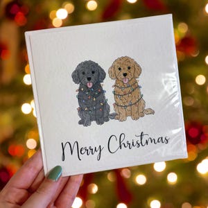 Peut inclure: Carte de Noël carrée avec deux chiens de dessin animé décorés de guirlandes lumineuses. Un chien est noir, l'autre est beige. Le texte "Merry Christmas" est écrit en dessous en cursive.